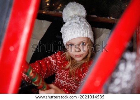 Blonde teenager girl in a white knitted hat with a pom-pom in a red car. Selective focus. Blurry lights.