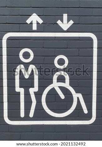 Closeup of isolated white elevator sign on grey wall with disabled person in wheelchair and campanion, up and down arrows