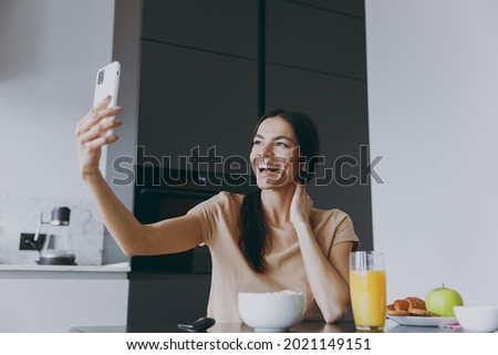 Young housewife woman in casual clothes beige t-shirt eat breakfast do selfie shot on mobile phone post photo on social network cook food in light kitchen at home alone Healthy diet lifestyle concept