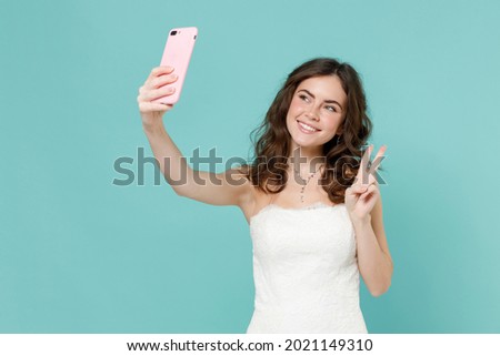 Smiling bride young woman 20s in white wedding dress doing selfie shot on mobile phone showing victory sign isolated on blue turquoise background studio portrait. Ceremony celebration party concept