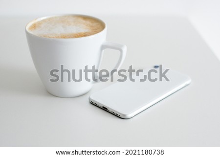 A white smartphone lies on a white table against a white wall. In the background a white cup with coffee. Minimalism. Minimalist interior. Clean home. Cleanliness. Composure. 
