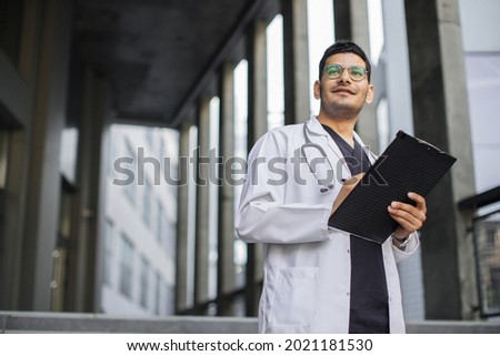Close up portrait of pensive professional male Arab doctor writing RX prescription on clipboard. Male doctor is filling in medical form standing outdoors of modern clinic. Copy space
