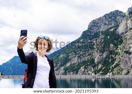 young adult woman taking a selfie in a mountain lake on a day hike, mountains in the background, horizontal