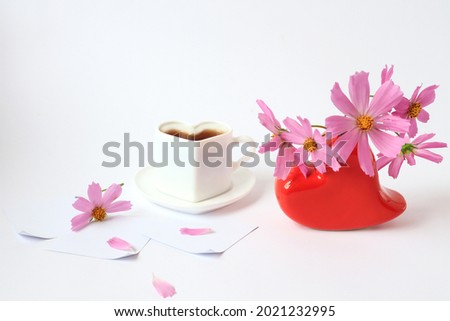 The concept of a good morning. Delicate pink flowers in a red heart-shaped vase, a cup of hot coffee on a light background, a light background, a side view, a place for text