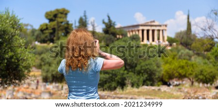 Girl tourist takes photo of Athens, Greece, Europe. Temple of Hephaestus, landmark of Athens in distance. Young pretty woman photographs attractions of Athens city. Travel and vacation in Greece.