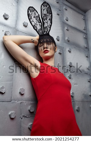 Young woman in black rabbit or hare fancy mask and red dress. Eyes closed. Hands up. In the background there is a metal wall with rivets .