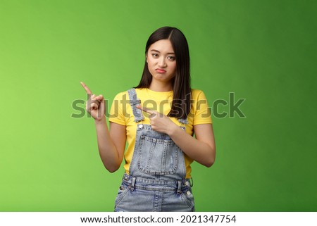 Disappointed uninterested asian gloomy girl pointing fingers left, grimacing sulking displeased, unwilling take part event, stare reluctant unimpressed, pose green background