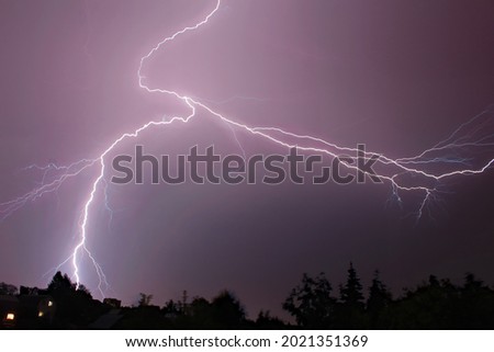 A huge forked lightning in the purple night sky. Bright flash in a thunderstorm, bad weather, a cataclysm. A low-angle shot of lightning silhouette above the houses. Concept: storm, hurricane, tornado