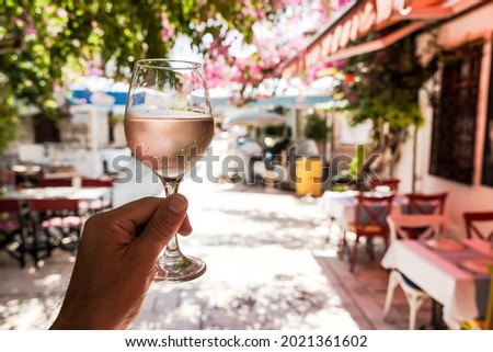 Glass of wine in hand. A glass of young fresh rose wine against the backdrop of a summer cafe in a Mediterranean seaside tourist town in the summer under sunlight. Summer, travel, lifestyle