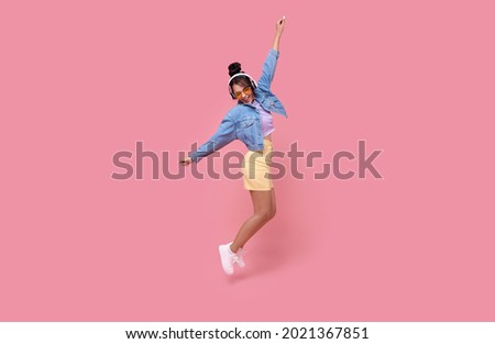 Happy beautiful Asian teen woman in yellow glasses listening music in headphones and dance on pink background.	