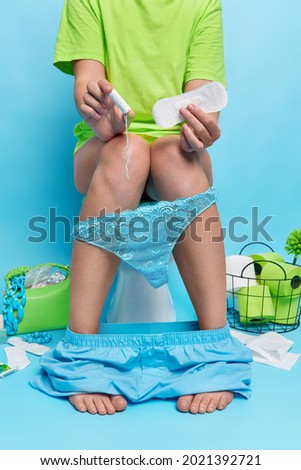 Faceless woman in casual clothes holds sanitary napkin and cotton tampon has menstruation poses on toilet bowl wears lacy pants pulled down on legs sits in restroom isolated over blue background.