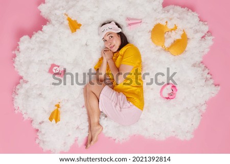 Overhead shot of dreamy Asian woman dressed in nightwear keeps hands together thinks about something pleasant before sleeping dreams about travel abroad lies on white fluffy cloud over rosy background