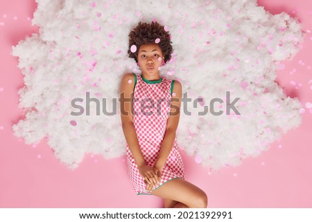 Romantic curly haired young woman keeps lips folded wants to kiss you dressed in checkered dress poses on white cloud with falling confetti on her has dreamy flirting expression pink background