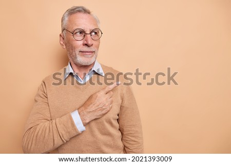 Horizontal shot of serious bearded elderly man points at upper right corner shows promo offer place for your advertising content wears round spectacles brown jumper poses indoor. Monochrome shot Royalty-Free Stock Photo #2021393009