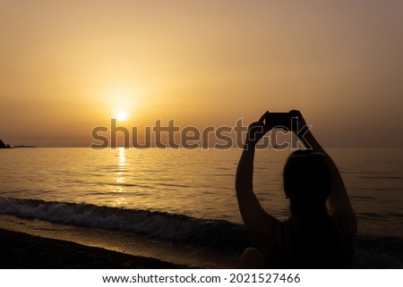 Silhouette of a woman taking photograph with her mobile phone at the seaside at dawn. Sun rises over the Mediterranean Sea on a lovely summer morning. Selective focus.