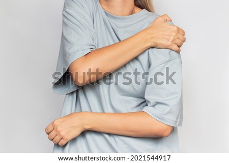 Cropped shot of a young woman in a gray t-shirt holding a shoulder in her hands isolated on a grey background. Shoulder injuries, arm pain, neuralgia Royalty-Free Stock Photo #2021544917