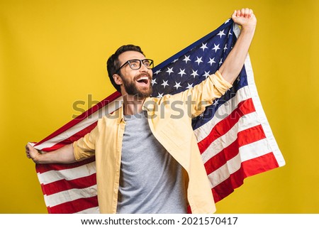 Portrait of young bearded happy man holding a USA american flag isolated over yellow background.