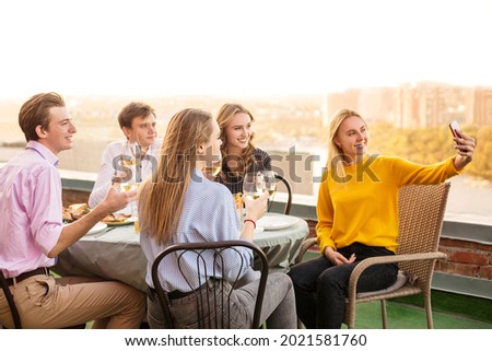 Group of five cheerful young people having dinner in restaurant, sitting on terrace by table and holding glasses of wine in hands posing for selfie on smartphone. Friendship and gathering concept