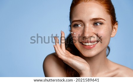Laughing redhead girl applying moisturizing cream on her face. Photo of young woman with flawless skin on blue background. Skin care and beauty concept