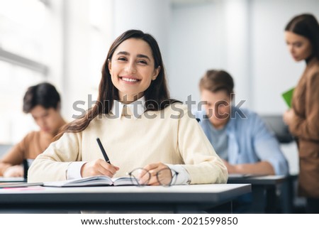 Modern Education Concept. Portrait of smiling female student sitting at desk in classroom at university, taking test or writing notes in her notebook, casual teen looking and posing at camera