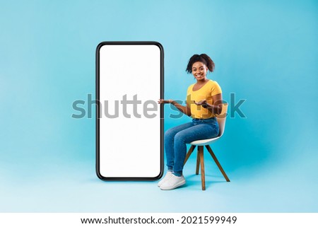 Full length of pretty Afro lady sitting on chair next to big blank smartphone with mockup for mobile app or website design, blue studio background. Space for your online advertisement Royalty-Free Stock Photo #2021599949