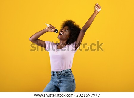 Music lover. Excited black woman singing songs in microphone and dancing, having fun and enjoying the moment, standing over yellow background, studio shot