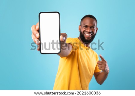 Excited black guy in casual wear pointing at cellphone with empty screen on blue studio background, space for website or mobile app design. Cellphone display mockup. Selective focus Royalty-Free Stock Photo #2021600036