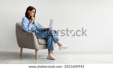 Full length of serious young woman looking at laptop screen, sitting in armchair, having problem with online work or studies against white studio wall, banner with copy space