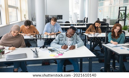 Modern Education Concept. Portrait of diverse group of students sitting at desks in classroom at university, taking test, entrance examination or writing notes in notebook, selective focus