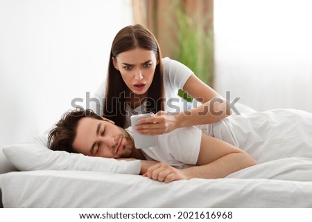 Infidelity. Shocked Wife Reading Message From Lover On Phone While Cheating Husband Sleeping In Bedroom At Home. Jealous Woman Checking Boyfriend's Cellphone Chats. Affair, Relationship Issues Royalty-Free Stock Photo #2021616968