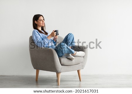 Positive young lady in casual wear sitting in armchair with crossed legs, warming hands on cup of hot coffee, enjoying warm drink against white studio wall, copy space Royalty-Free Stock Photo #2021617031