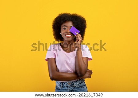 Purchase, money and payment concept. Joyful african american woman covering eye with credit card, wearing casual clothes posing isolated on yellow wall background, studio shot. Royalty-Free Stock Photo #2021617049