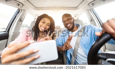 Beautiful young African American couple taking selfie inside car, using mobile phone, showing v victory peace sign gesture. Happy black man and woman going on vacation, taking self photo in auto