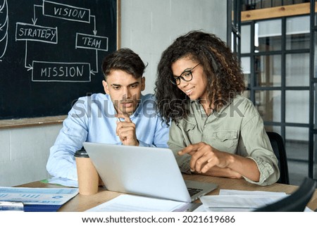 Young smiling African American mentor explaining to serious Indian coworker project strategy. Diverse startup coworkers students girl and guy talking discussing working in modern office using laptop. Royalty-Free Stock Photo #2021619686