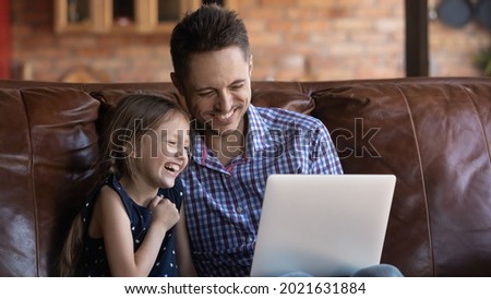 Happy daughter girl and dad watching funny comedy movie on laptop at home, sitting on couch, enjoying entertainment together, talking to family or mom via video call, looking at screen laughing