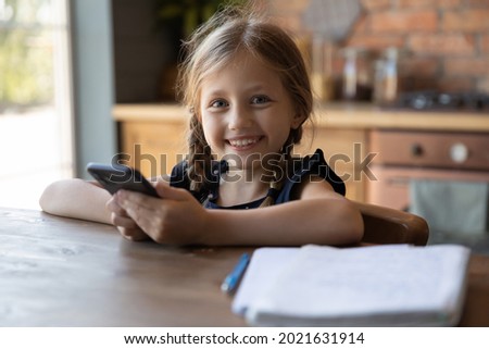 Portrait of happy gen Z preteen school pupil girl holding smartphone, looking at camera, smiling. Sweet schoolkid with gadget learning on internet from home, using mobile phone for studying, Head shot