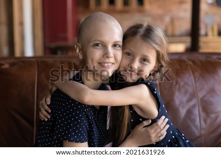 Portrait of happy optimistic cancer patient mom embracing little daughter on couch, enjoying leisure with family, spending time at home. Cute affectionate girl giving hug and support to sick mom