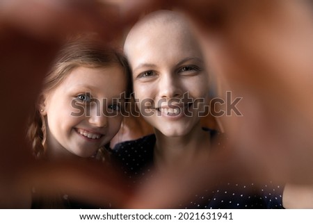 Happy young mom with cancer and cute little daughter kid having fun, smiling at camera through heart shaped hand frame. Optimistic patient enjoying leisure time with child. Close up portrait