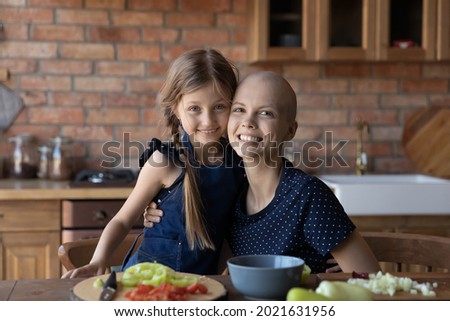Happy little kid and hairless mom cooking lunch in kitchen, hugging, looking at camera, smiling. Cancer mother and loving daughter enjoying home activity, preparing salad together. Head shot portrait