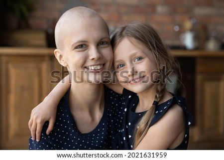 Happy cancer mom and lute little daughter hugging at home, looking at camera, smiling, enjoying leisure time. Kid giving love, support to ill hairless mother. Oncology in family. Headshot portrait