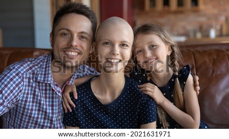 Happy optimistic hairless young woman with cancer feeling family support, love, care. Headshot portrait of parents and little kid sitting on couch at home, hugging, looking at camera, smiling