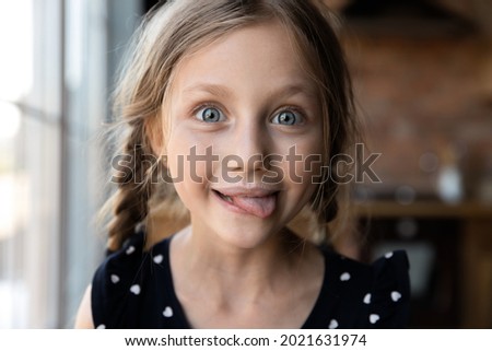 Happy funny kid with white moustache showing tongue, licking milk from upper lip, looking at camera. Girl drinking milk, enjoying yummy dairy product, source of calcium and protein. Head shot portrait