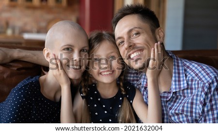 Happy family sitting on couch at home together, looking at camera, smiling. Cute little daughter girl touching faces of dad and hairless mom. Young cancer patient getting support from husband and kid
