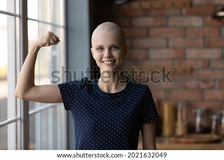 Happy strong cancer survivor winning fight for life, beating disease. Young woman with shaved head showing strength, hand muscles power, flexing bicep, looking at camera, smiling. Head shot portrait