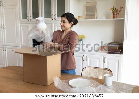 Happy young Indian female renter or tenant settle in new home unbox parcel shopping tableware online from home. Smiling ethnic woman unpack box buying kitchenware on internet. Delivery concept. Royalty-Free Stock Photo #2021632457