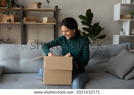 Smiling woman unpacking parcel, excited young female opening cardboard box, sitting on cozy couch at home, satisfied curious customer received online store order, good delivery service concept Royalty-Free Stock Photo #2021632571