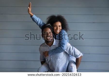 Overjoyed young African American couple renters have fun hug cuddle isolated on grey wall background. Smiling millennial ethnic woman piggyback happy biracial man show love. Rental, moving concept.