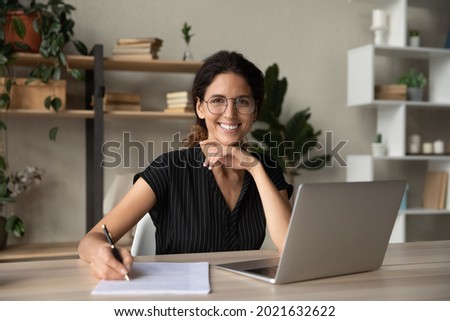 Head shot portrait of smiling woman in glasses writing, taking notes, female student sitting at work desk with laptop, studying online, webinar, confident businesswoman working with documents Royalty-Free Stock Photo #2021632622