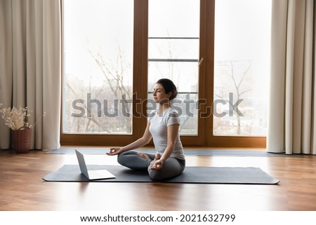 Calm millennial Indian woman sit on mat at home meditate watch online training or session on laptop. Peaceful young ethnic female practice yoga breathe fresh air have webcam call with trainer. Royalty-Free Stock Photo #2021632799