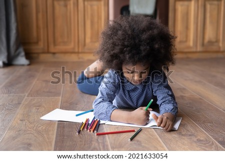 Cute small teen African American girl child lying on warm wooden kitchen floor drawing painting in album alone. Little teenage ethnic biracial kid have fun rest at home engaged in art hobby activity.